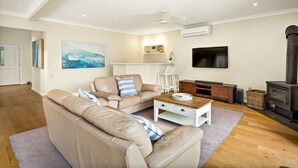 A living room with a beige sofa, a coffee table, a TV, a fireplace, and a bookshelf. - Sandy Feet Retreat in Cape Woolamai (Cape Woolamai)