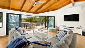 A modern living room with a large sofa, a coffee table, and a TV. There is a balcony with outdoor furniture and a view of trees. - Sandy Feet Retreat in Cape Woolamai (Cape Woolamai)