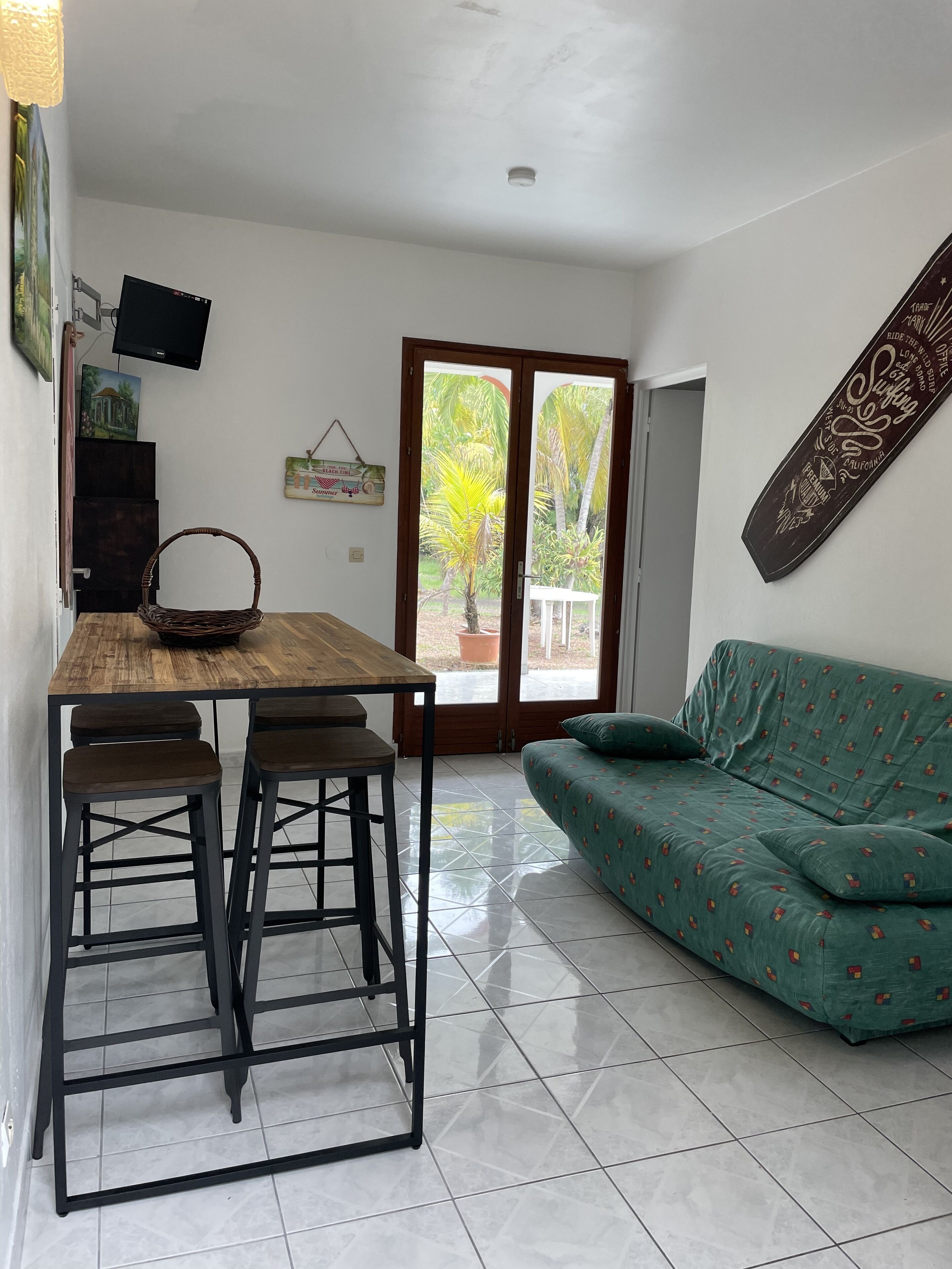 Una habitación con una mesa de comedor de madera, dos taburetes altos, un sofá verde y un ventanal corredero que da a un espacio exterior con plantas.