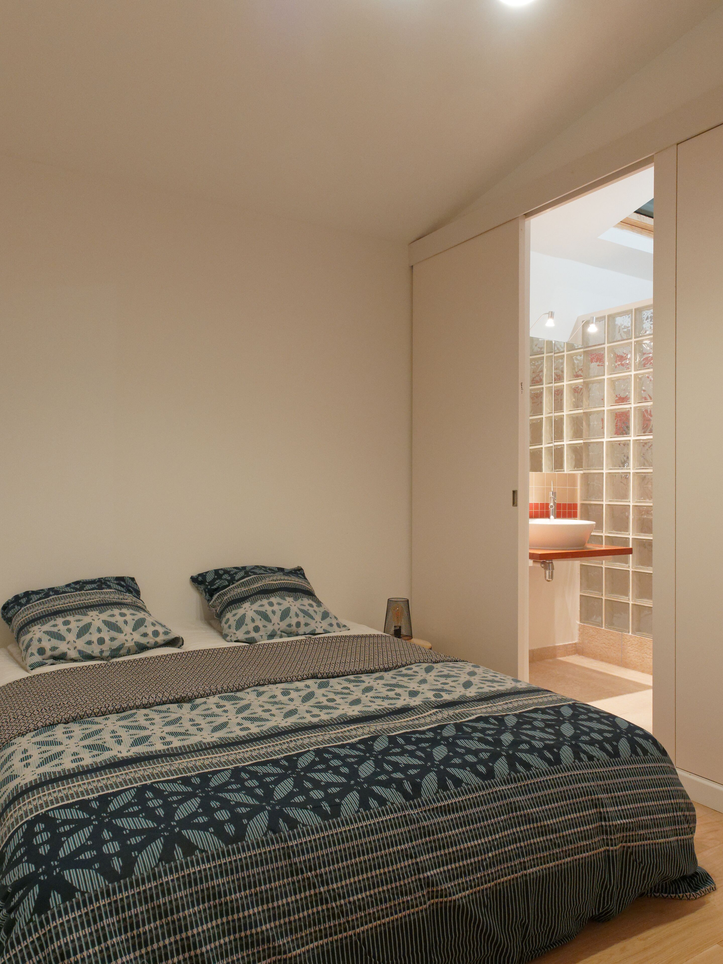 Un dormitorio con cama, dos almohadas, una mesita de noche y vistas a un baño por una puerta abierta.