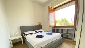 Ein ordentlich gemachtes Bett mit Kissen und gefalteten Handtüchern, ein Nachttisch mit Lampe sowie ein Fenster mit Blick auf Grünflächen.
