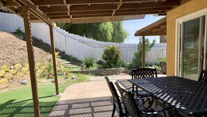 Un patio couvert avec une table et des chaises, une pergola et une vue sur un jardin agrémenté d’une clôture blanche et d’arbres.