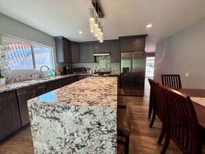 Una cocina moderna con armarios oscuros, una isla grande de mármol, electrodomésticos de acero inoxidable y una zona de comedor con sillas de madera.