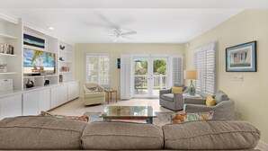 Ein geräumiges Wohnzimmer mit einer großen Couch, einem Couchtisch und einem Fernseher auf einer weißen Unterhaltungsanlage. Ein Deckenventilator, eine Glasschiebetür zu einer Außenpatio und ein gerahmtes Bild an der Wand sind ebenfalls vorhanden.