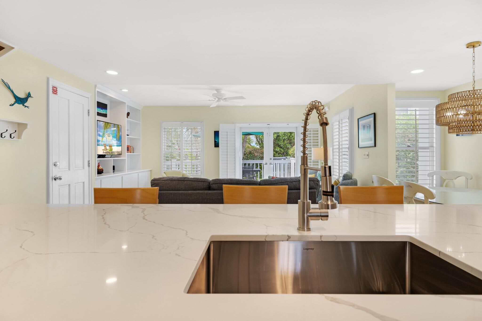 A modern kitchen with a marble countertop, stainless steel sink, and a built-in oven. There is a dining area with a white table and chairs, and a living room with a dark sofa and a white coffee table.