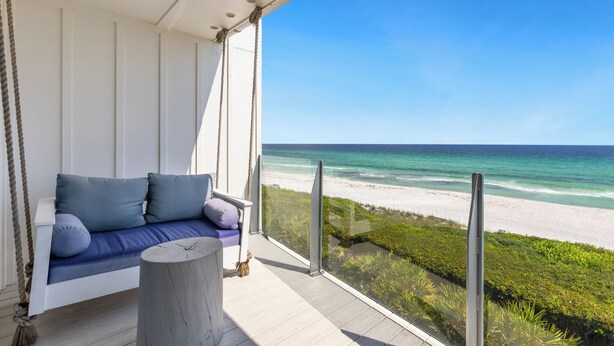 A balcony with a white swing seat and a round wooden table, overlooking a beach and clear blue sky.
