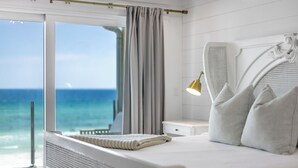 A bedroom with a large window, a bed with pillows, a bedside lamp, and a view of the ocean.