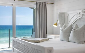 A bedroom with a large window, a bed with pillows, a bedside lamp, and a view of the ocean.