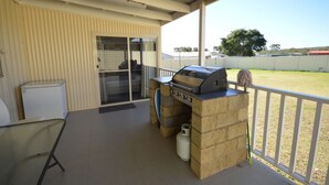 Une terrasse couverte avec un barbecue, une porte vitrée et une clôture blanche en arrière-plan.