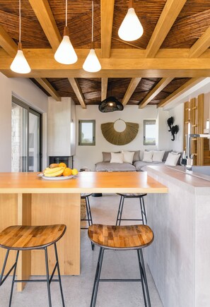 A modern kitchen with a wooden island, bar stools, hanging pendant lights, and a fruit bowl. - Allure Retreat Villa (Λευκάδα)