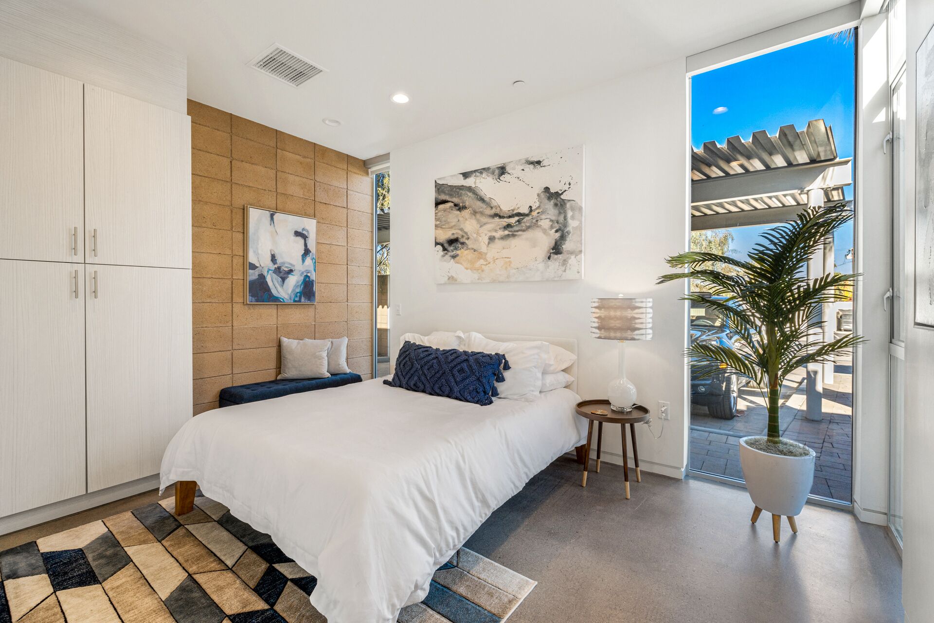 A modern bedroom with a large bed, a wooden accent wall, abstract art, a potted plant, and a view of the outdoors.