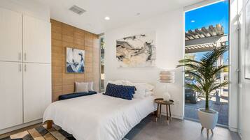 A modern bedroom with a large bed, a wooden accent wall, abstract art, a potted plant, and a view of the outdoors.
