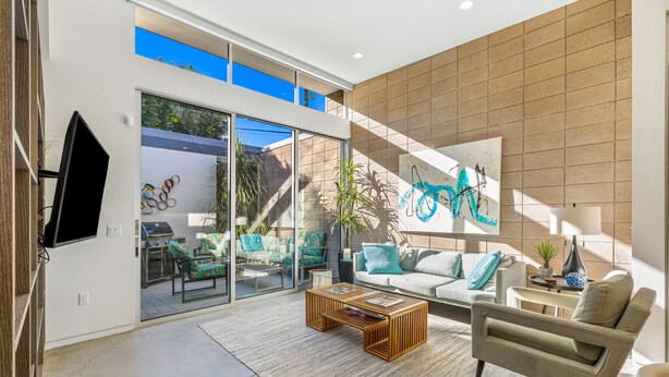 A modern living room with a large sliding glass door leading to a patio, a flat-screen TV mounted on the wall, and a contemporary art piece above the sofa. - Palm Desert Velo - Modern Condo w Spa (Palm Desert)
