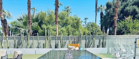 A pool with clear water, surrounded by palm trees and cacti, with lounge chairs and a sunbed in the background.