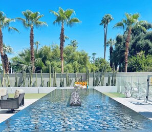 A pool with clear water, surrounded by palm trees and cacti, with lounge chairs and a sunbed in the background.
