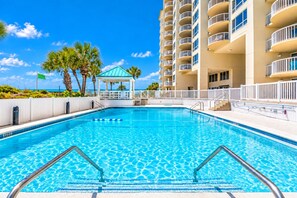 A swimming pool with clear blue water, surrounded by a white fence, and a building with multiple balconies in the background. - Gulfside 505 - Gulf Front, Community Pool, Hot Tub, Tennis, Basketball, Fitness! (Miramar Beach)
