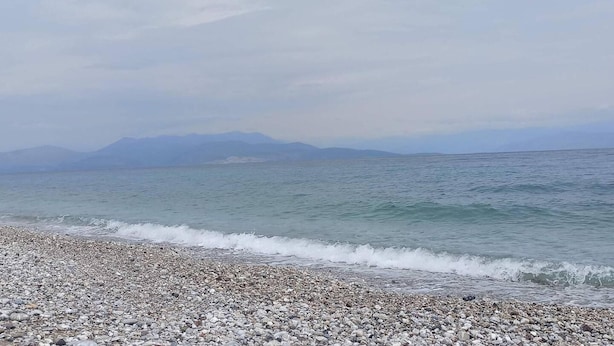 Ein Kiesstrand mit sanften Wellen, eine entfernte Bergkette und ein klarer Himmel.
