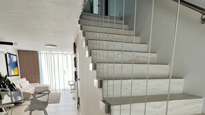 A modern staircase with a glass railing, a minimalist living area with a sofa and a coffee table, and a view of the outdoors through large windows. - Dream Villa · Direct Beach Access · Pool · 2BR Gated · Dorado, PR (Dorado)