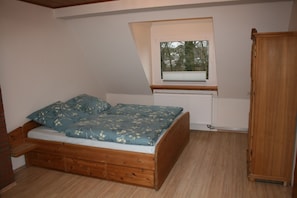 A small bedroom with a loft bed, a window with blinds, a radiator, and a wooden wardrobe.