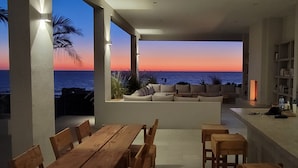 Un espace de vie moderne avec vue sur l’océan au coucher du soleil, comprenant une table en bois, des sièges et une bibliothèque.