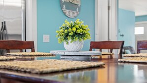 Uma mesa de jantar com um vaso branco cheio de flores verdes no centro, rodeada por individuais de palha e cadeiras de madeira.