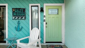 Uma casa turquesa com porta verde, uma cadeira branca e uma placa com a inscrição 'SALTY BREEZES'.