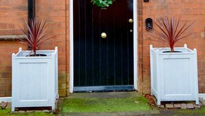 A black door with a green wreath, flanked by two white planters with red plants, and a brick wall with a black pipe. - Remarkable 1 Bed Apartment in Birmingham (Birmingham)