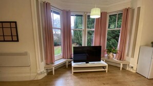 A room with a TV on a white stand, large windows with pink curtains, a white radiator, and a small white cabinet. - Remarkable 1 Bed Apartment in Birmingham (Birmingham)