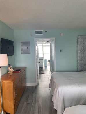 Um quarto com cama, penteadeira, abajur e televisão na parede.