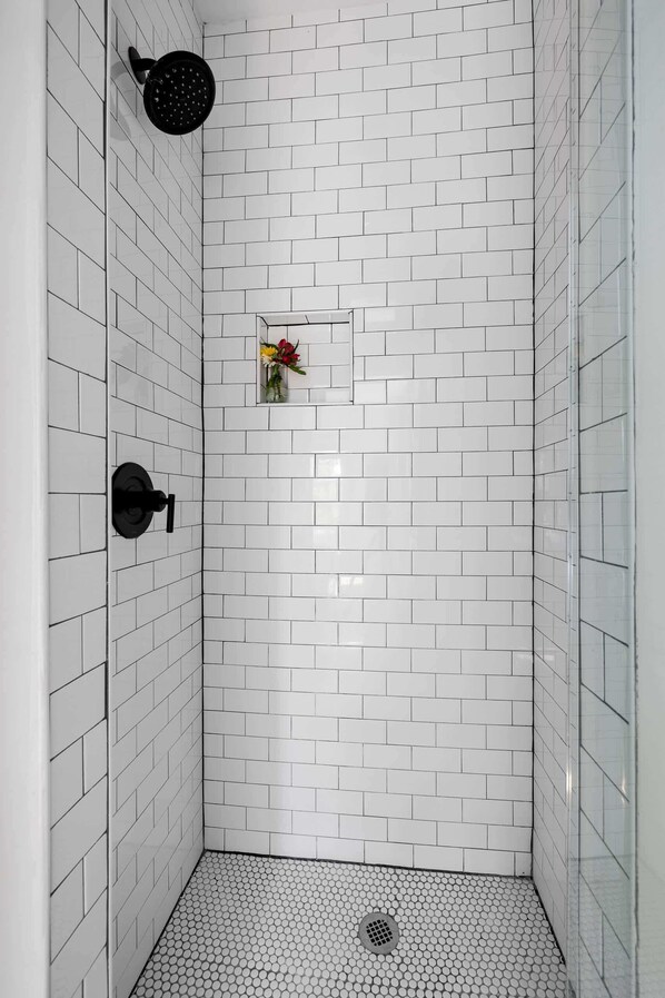 Ducha con paredes y suelo de azulejos blancos, cabezal de ducha negro y un pequeño jarrón con flores en la pared.