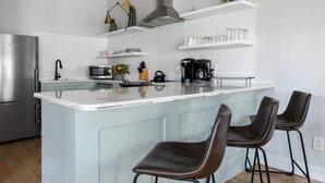 Una cocina moderna con una isla central, taburetes de bar, un refrigerador de acero inoxidable, microondas, cafetera y una planta colgante.