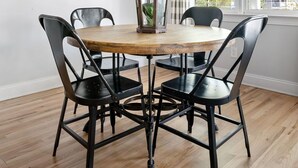 Un comedor con una mesa redonda de madera, sillas negras, una lámpara colgante de fibras tejidas, un cuadro abstracto en la pared y una ventana con persianas de bambú.