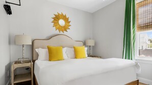 Une chambre d’hôtel avec un lit, deux tables de chevet, une lampe sur chacune, un miroir rond au mur et une fenêtre avec des rideaux verts.