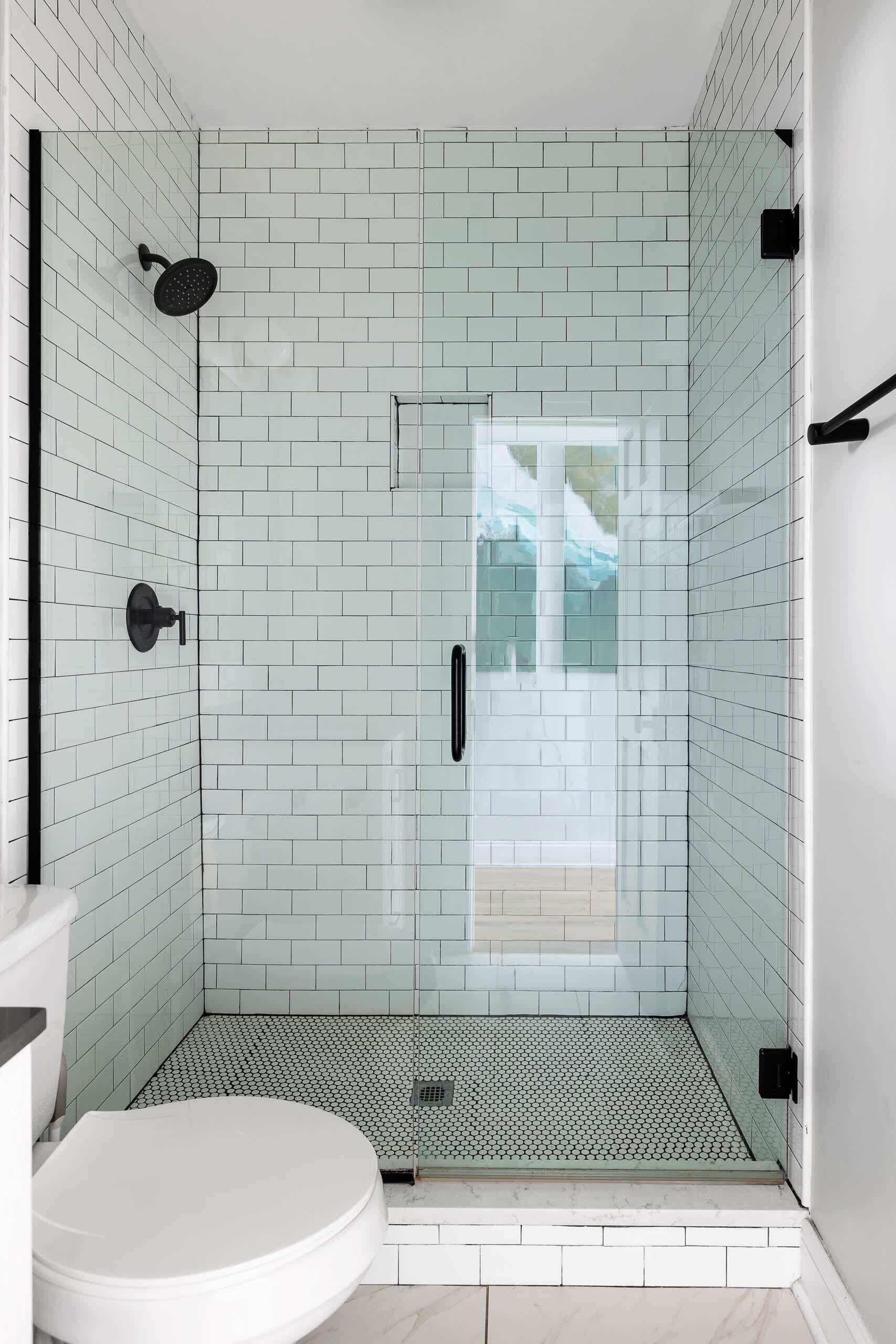 Une douche avec une porte en verre, des murs recouverts de carreaux blancs et des toilettes blanches dans un coin.