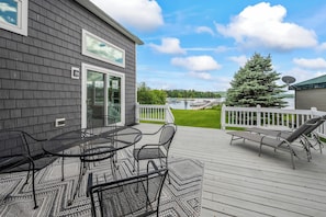 A deck with a table and chairs, a white fence, and a view of a body of water and trees.