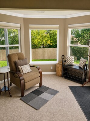 A corner room with a brown armchair, a bench with cushions, a small table, and a view of a backyard through large windows. - Private Hot Tub & Sunset Views - 3 BR Guest Suite (North Salt Lake)