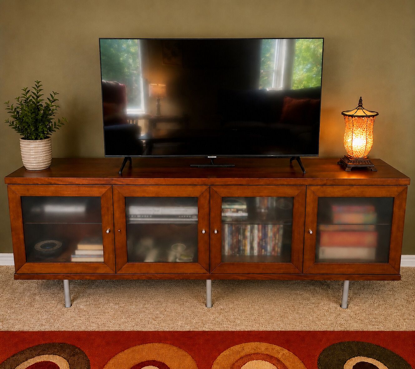 A flat-screen TV on a wooden cabinet with glass-fronted storage compartments, a potted plant, and a decorative lamp.