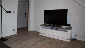 Una sala moderna con un televisor de pantalla plana sobre un mueble blanco, piso de madera y una puerta que da a otra habitación.