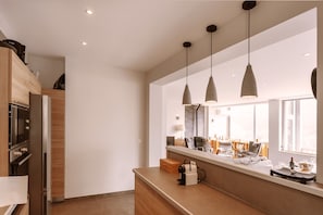 A modern kitchen with wooden cabinets, a built-in oven, and a long countertop. There are pendant lights above the counter and a window providing natural light. - Experience the Exception at Chalet La Source: Comfort, Relaxation and Magnificent Views (Cauterets)