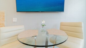Un tavolo rotondo in vetro con due sedie color crema, una piccola pianta in vaso e un televisore che mostra una vista dell'oceano blu.
