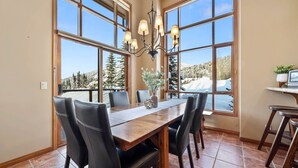 Ein Essbereich mit Holztisch, schwarzen Stühlen, einer Chandelierleuchtre und großen Fenstern mit Blick auf schneebedeckte Bäume.