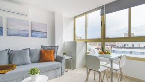 Una sala moderna con un sofá gris, una pequeña mesa blanca con una planta y un comedor con una mesa de vidrio y sillas blancas.