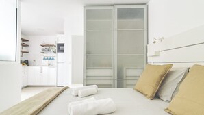 Una habitación de hotel moderna con una cama grande, una puerta corrediza de vidrio a un balcón y una kitchenette al fondo.