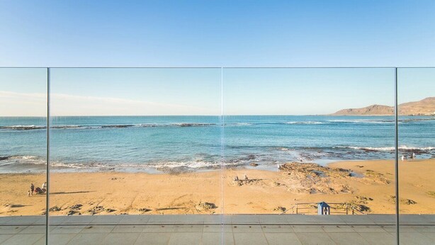 Vista a la playa a través de un panel de vidrio, con suelo de baldosas, playa arenosa y océano.