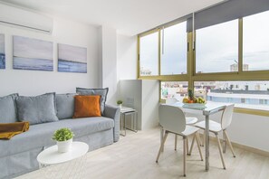 Una sala moderna con un sofá gris, una mesita blanca con una planta y una zona de comedor con una mesa de vidrio y sillas.