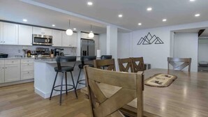 Una cuina moderna amb armaris blancs, una illa central amb tamborets, una taula de dinar amb cadires i un llum penjant a la paret.