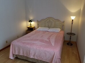 A neatly made bed with a pink blanket, a bedside table with a lamp, and a floor lamp in a simple room with wooden flooring.
