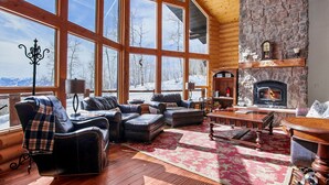 A log cabin interior with large windows, a stone fireplace, leather furniture, and a view of a snowy landscape. - Luxurious Mountain Retreat - Minutes from Park City and Deer Valley (Coalville)