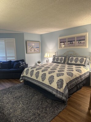 A bedroom with a bed, a nightstand, a lamp, a framed picture, and a window with blinds. - Oceanside vacation condo (Wildwood Crest)
