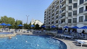 Ein Hotelpoolbereich mit Liegestühlen und Sonnenschirmen, umgeben von einem mehrstöckigen Gebäude mit Balkonen.